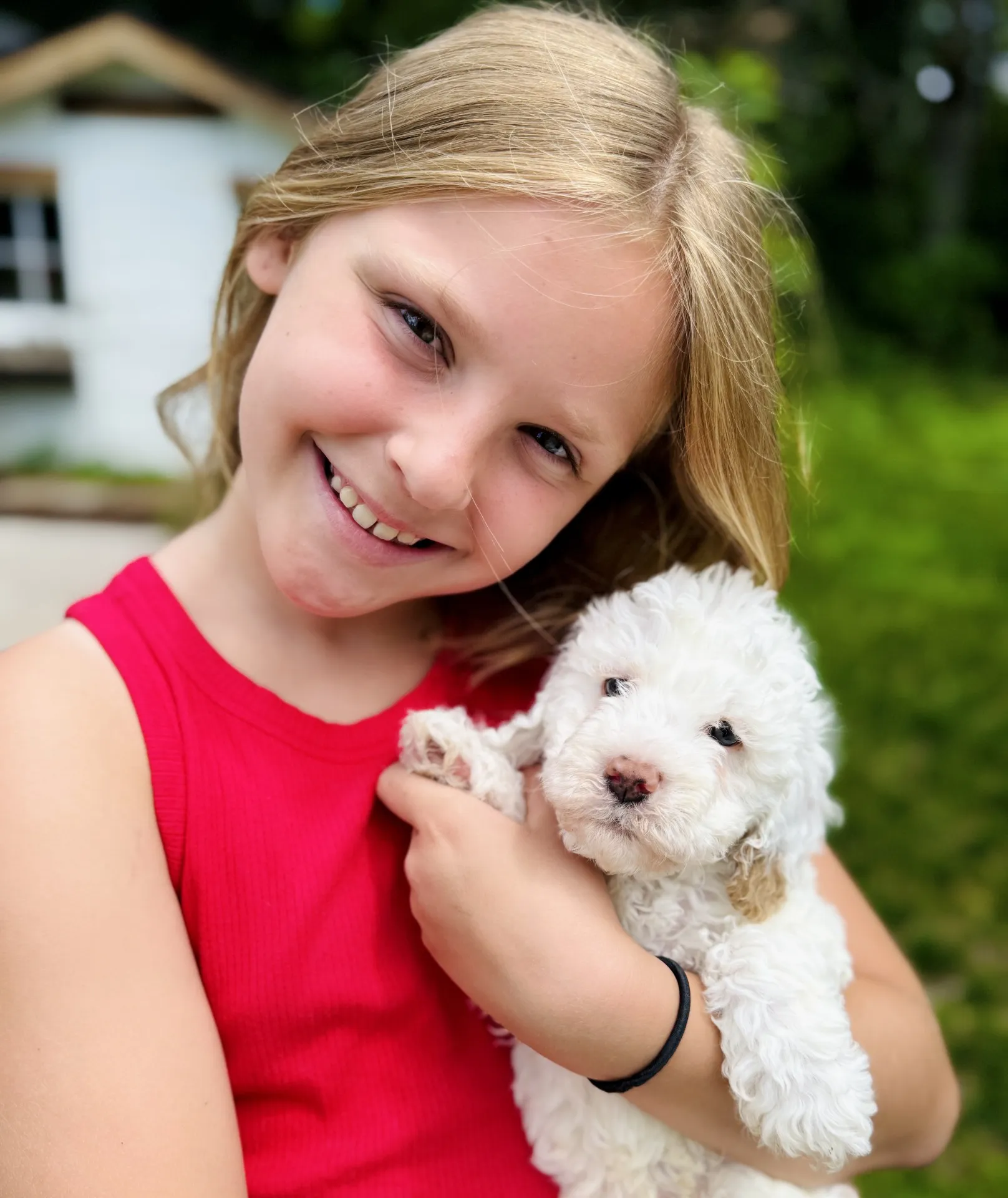HPF - A smiling child in a red shirt holds a fluffy white puppy outside, with greenery and a house visible in the background. - Happy Farm Puppies